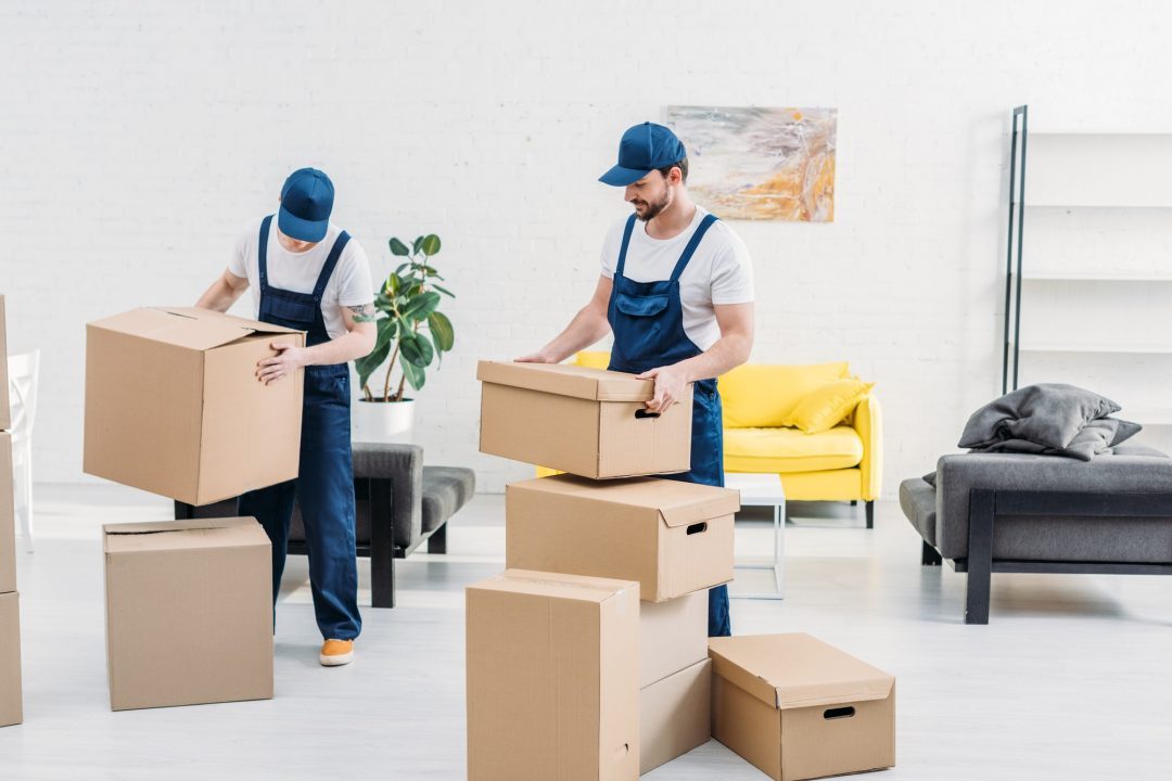 two-movers-in-uniform-carrying-cardboard-boxes-in-modern-apartment-1.jpg